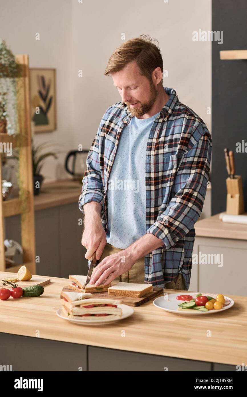 Young man preparing sandwiches with vegetables at wooden table in kitchen Stock Photo