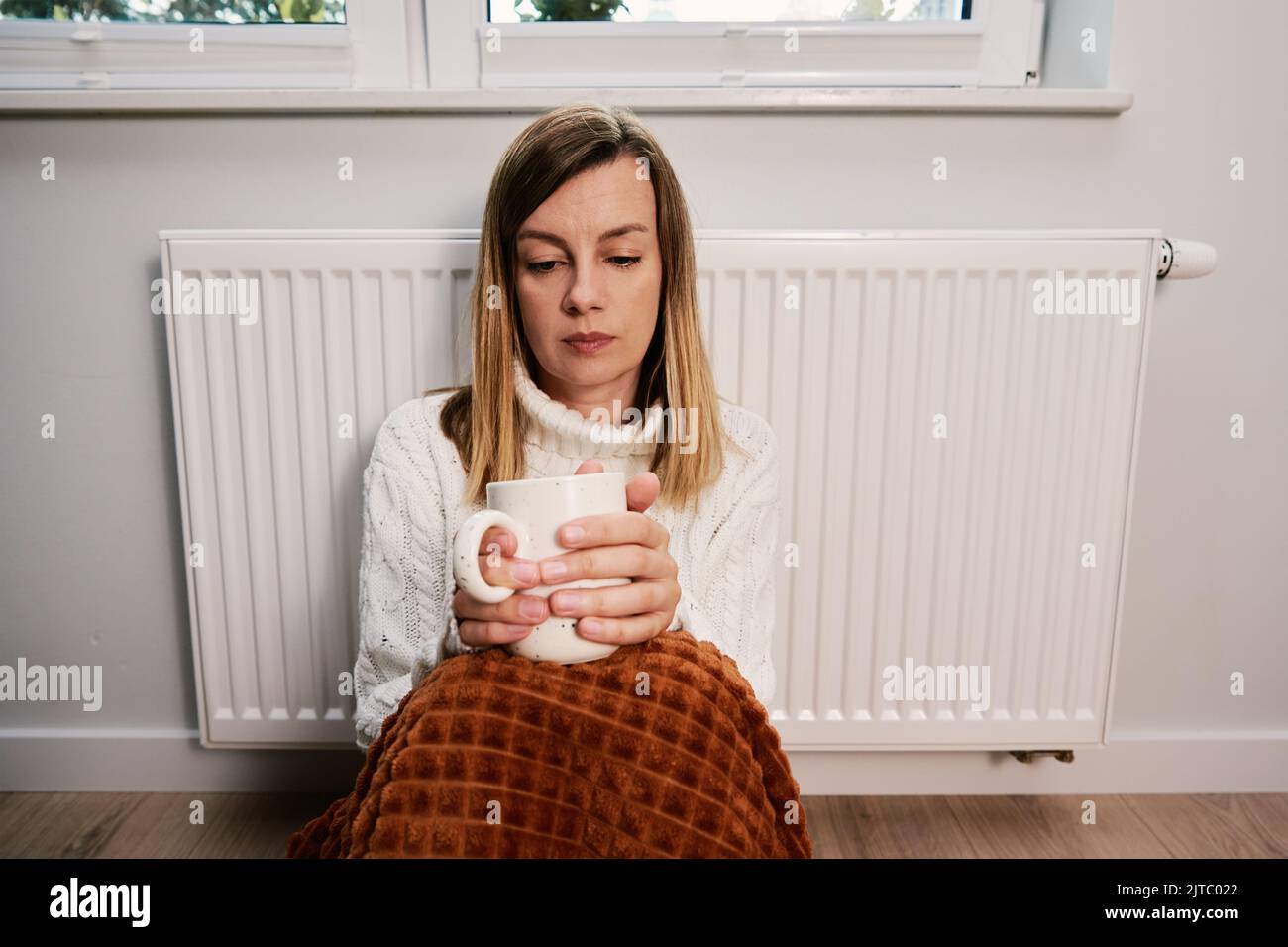 Worried sad woman sits under blanket near heating radiator with cup of tea, Rising costs in private households for gas bill due to inflation and war, Energy crisis Stock Photo