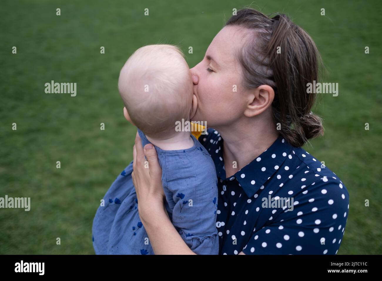Young mom holding and kissing baby outdoor on green grass background Stock Photo