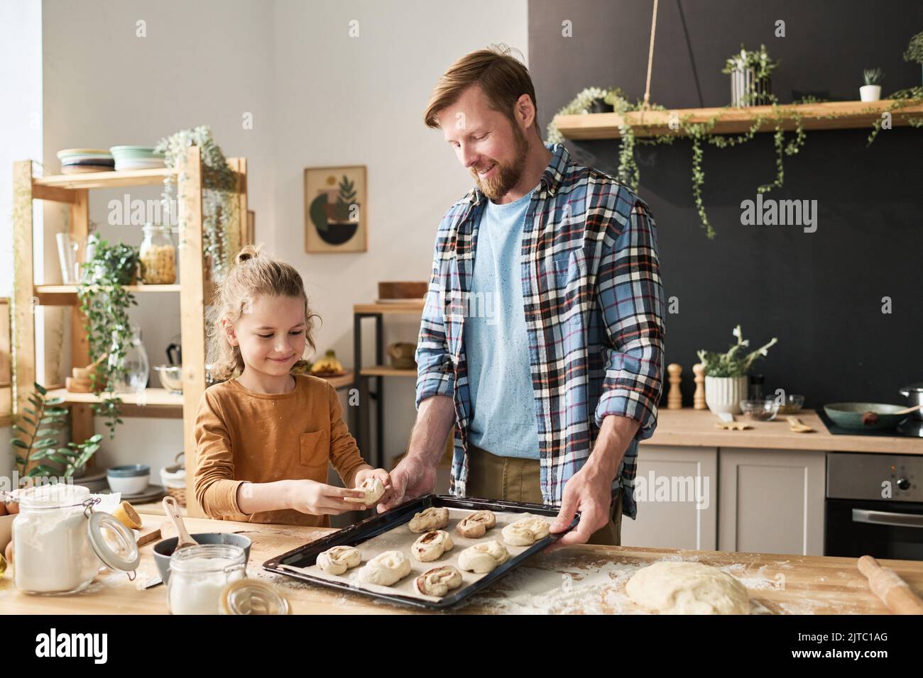 Little son putting homemade buns on tray for baking together with his dad in kitchen Stock Photo