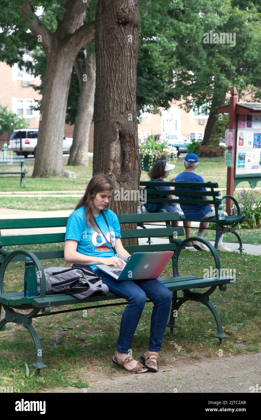 Young adult female using Apple laptop computer while sitting on a park bench. Stock Photo