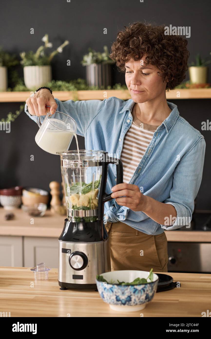 Young woman adding milk in blender to prepare fruit smoothie at table in kitchen Stock Photo
