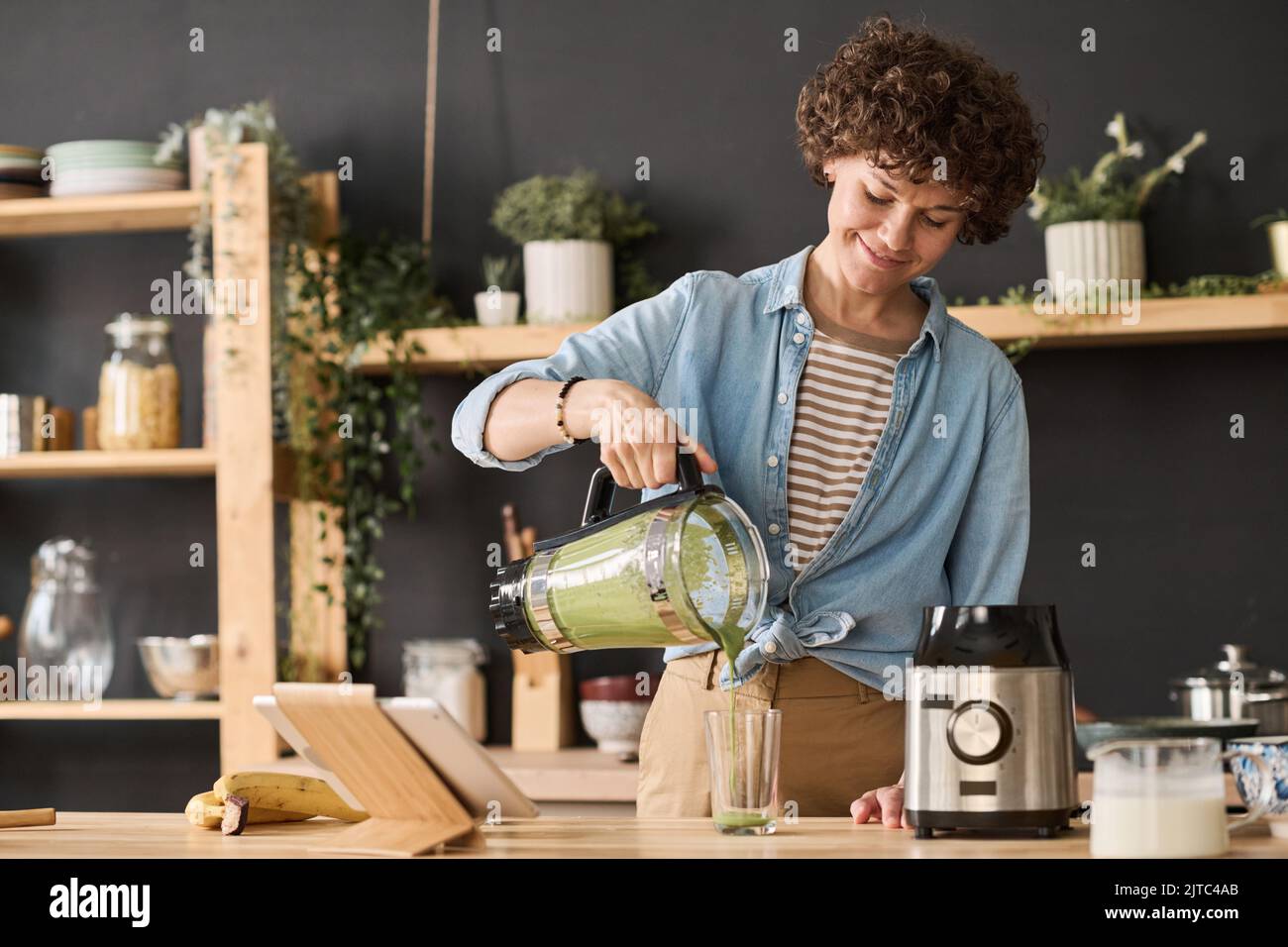 Young woman pouring fresh smoothie from blender in glass while standing at table in kitchen Stock Photo