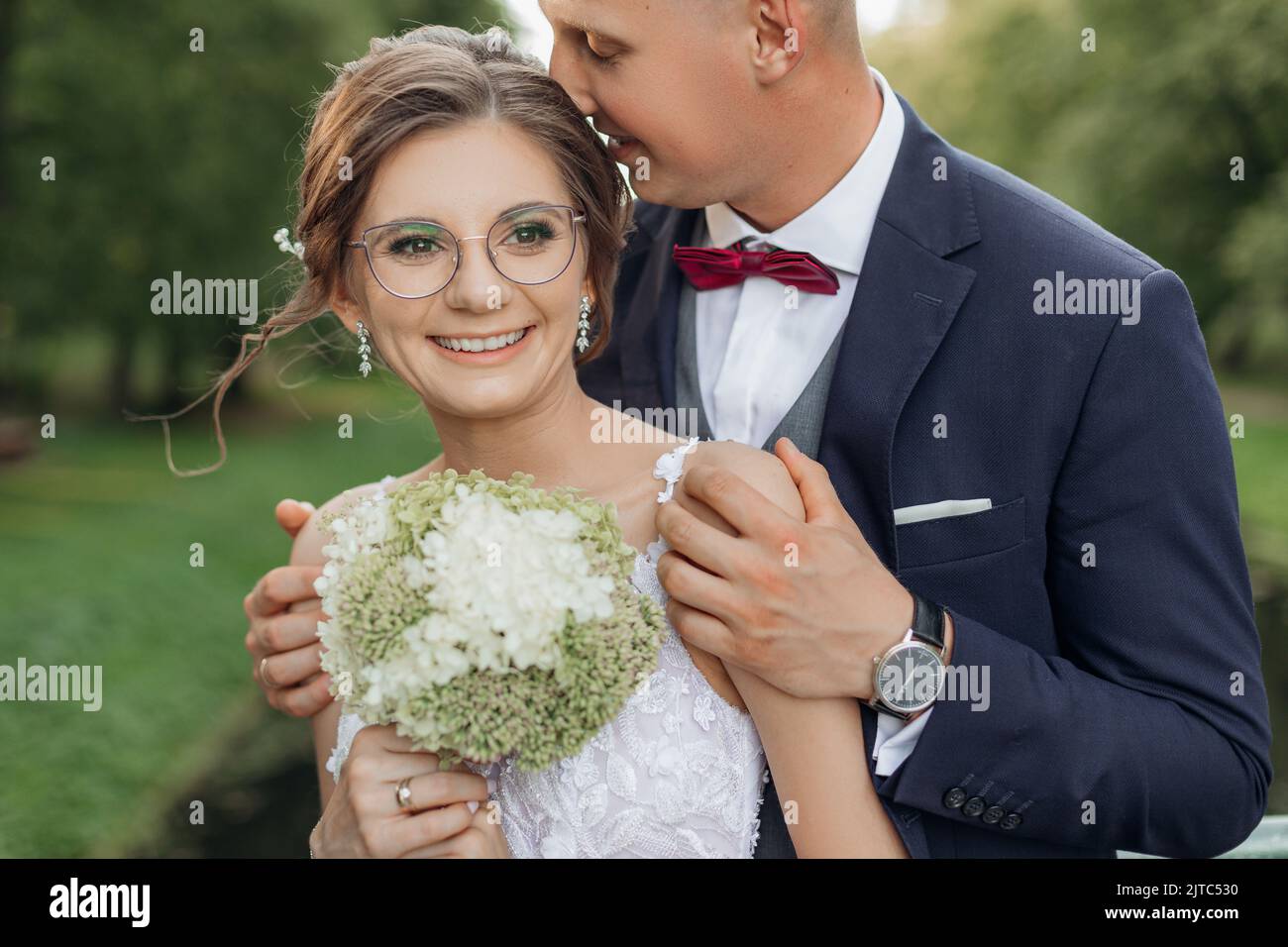 Charming, excited married couple of groom kissing bride in glasses with bouquet of flowers, hugging and stand together Stock Photo