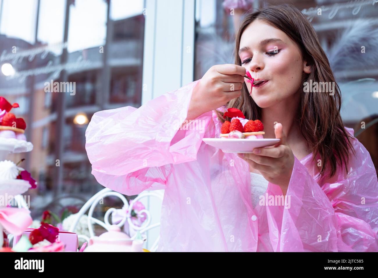 Pretty and calm teenage girl in pink raincoat joying, tasting sweet raspberry dessert cakes in outdoor cafe near window Stock Photo