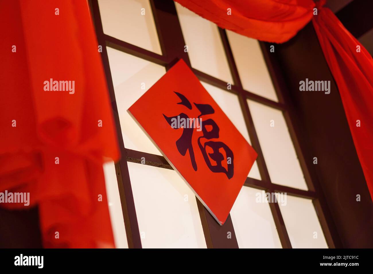 A low angle shot of Chinese calligraphy written on a square board hanging on a closed window with red fabric surrounding the window Translation: luck Stock Photo