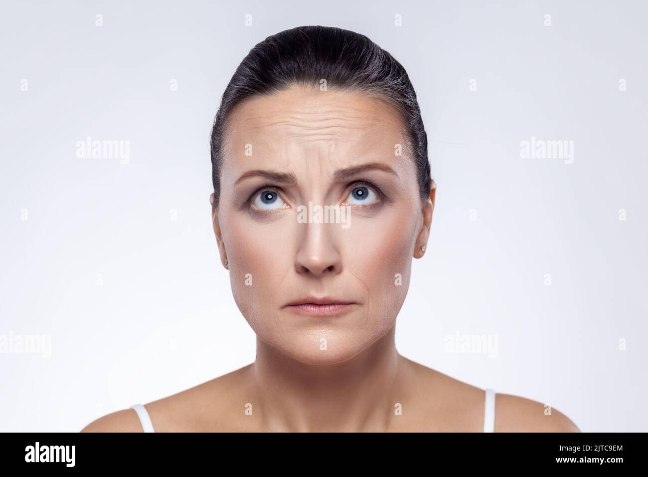 Closeup portrait of well-groomed middle aged woman checking wrinkles on her forehead, looking up, anti aging rejuvenation, skin care. Indoor studio shot isolated on white background. Stock Photo