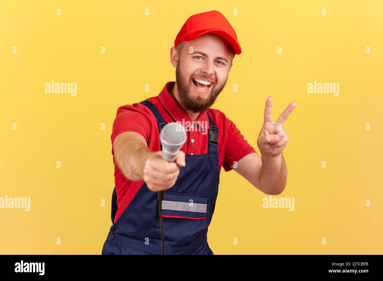 Portrait of positive bearded worker man wearing blue uniform and red cap offering microphone for answering questions and showing v sign. Indoor studio shot isolated on yellow background. Stock Photo