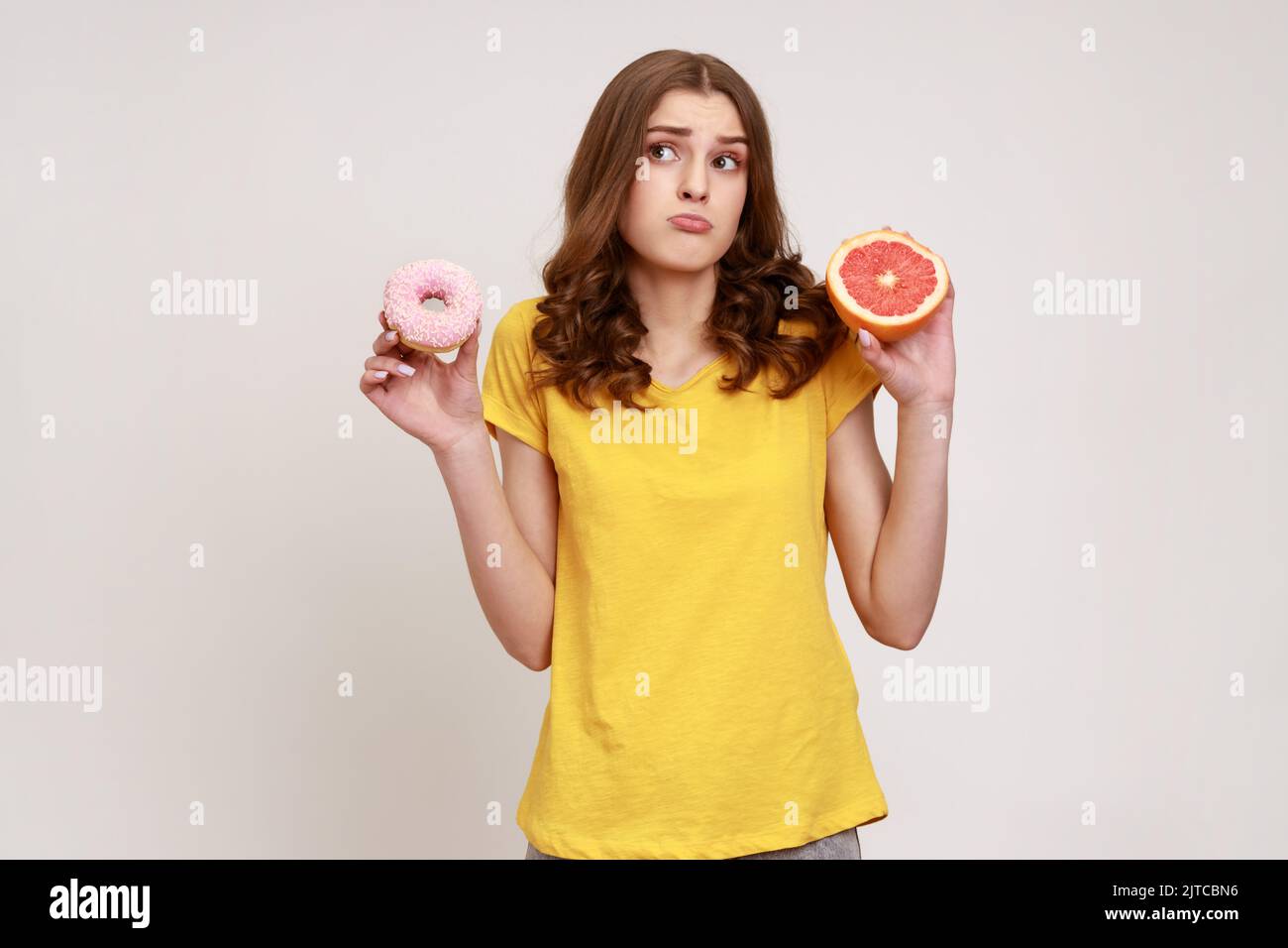 Healthy fruits vs high-calorie dessert. Portrait of confused curly-haired teen girl in casual style T-shirt choosing between grapefruit and donut. Indoor studio shot isolated on gray background. Stock Photo