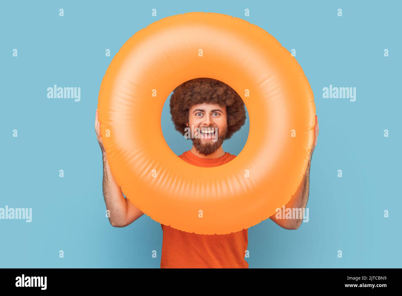 Portrait of man with Afro hairstyle with happy facial expression looking trough bright orange rubber ring, looking at camera, enjoying his vacation. Indoor studio shot isolated on blue background. Stock Photo