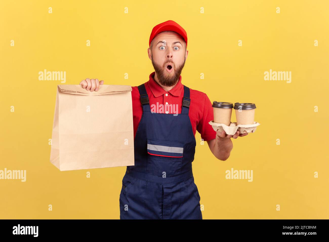 Portrait of astonished barded deliveryman wearing uniform holding paper package and take away coffee in hands, fast delivery, breakfast. Indoor studio shot isolated on yellow background. Stock Photo