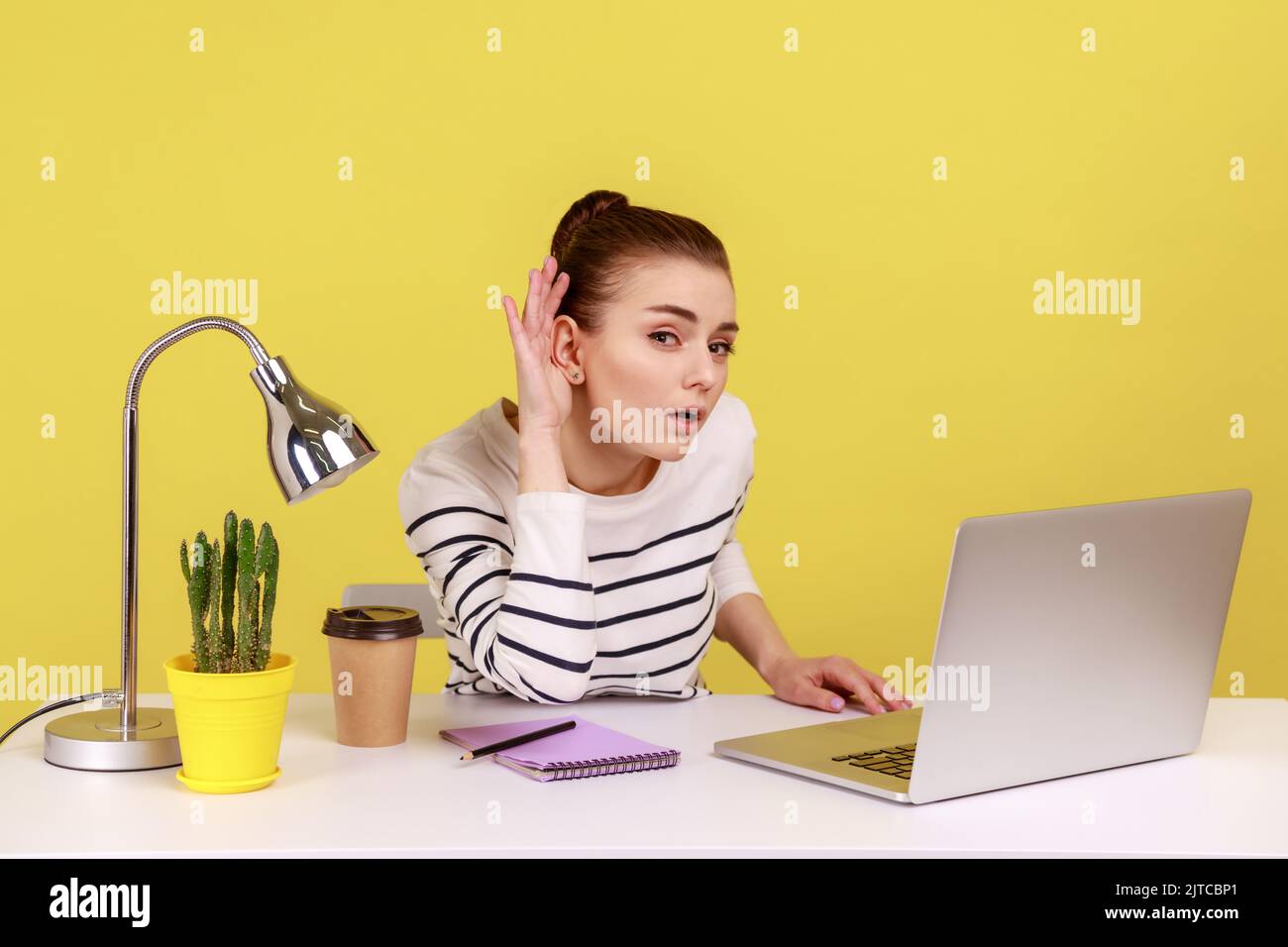 I can't hear you. Woman manager holding arm near ear trying to listen secret while working online on laptop in home office. Indoor studio studio shot isolated on yellow background. Stock Photo