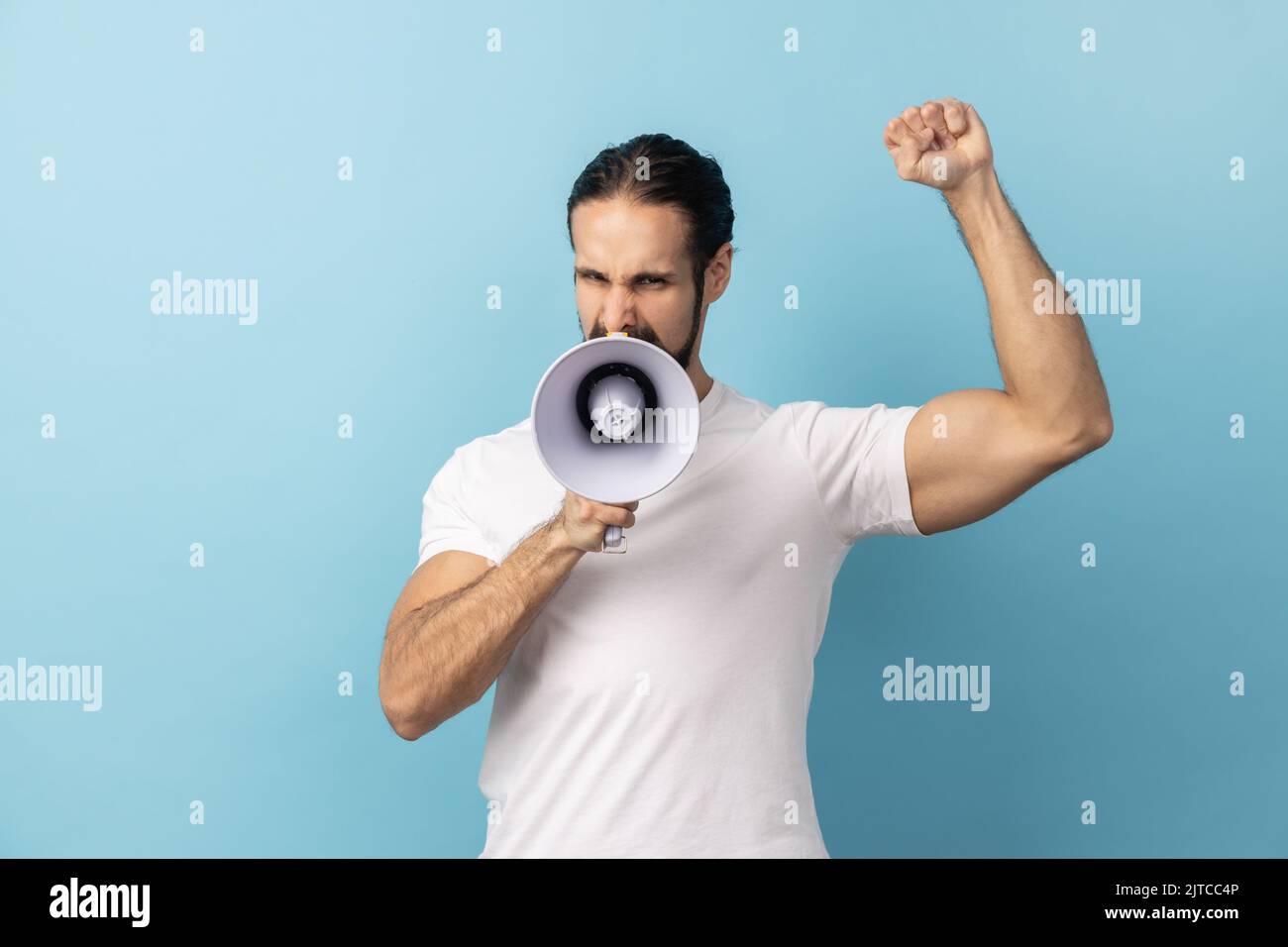 Portrait of angry man with beard wearing white T-shirt loudly screaming at megaphone, making announce, protesting, wants to be heard. Indoor studio shot isolated on blue background. Stock Photo