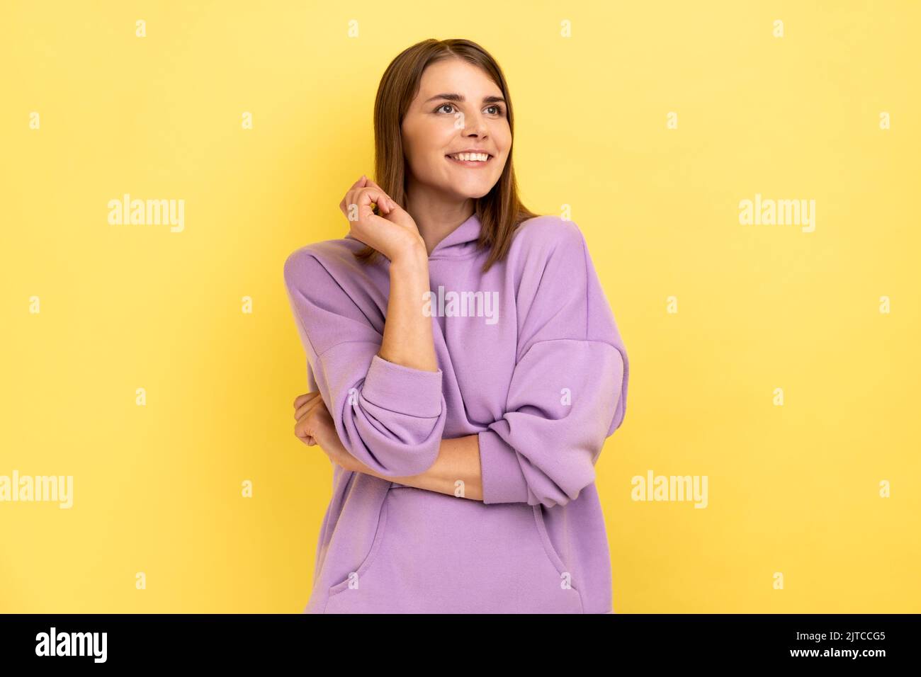 Portrait of adorable dreamy young woman standing and dreaming, making wish, love, looking away with toothy smile, wearing purple hoodie. Indoor studio shot isolated on yellow background. Stock Photo