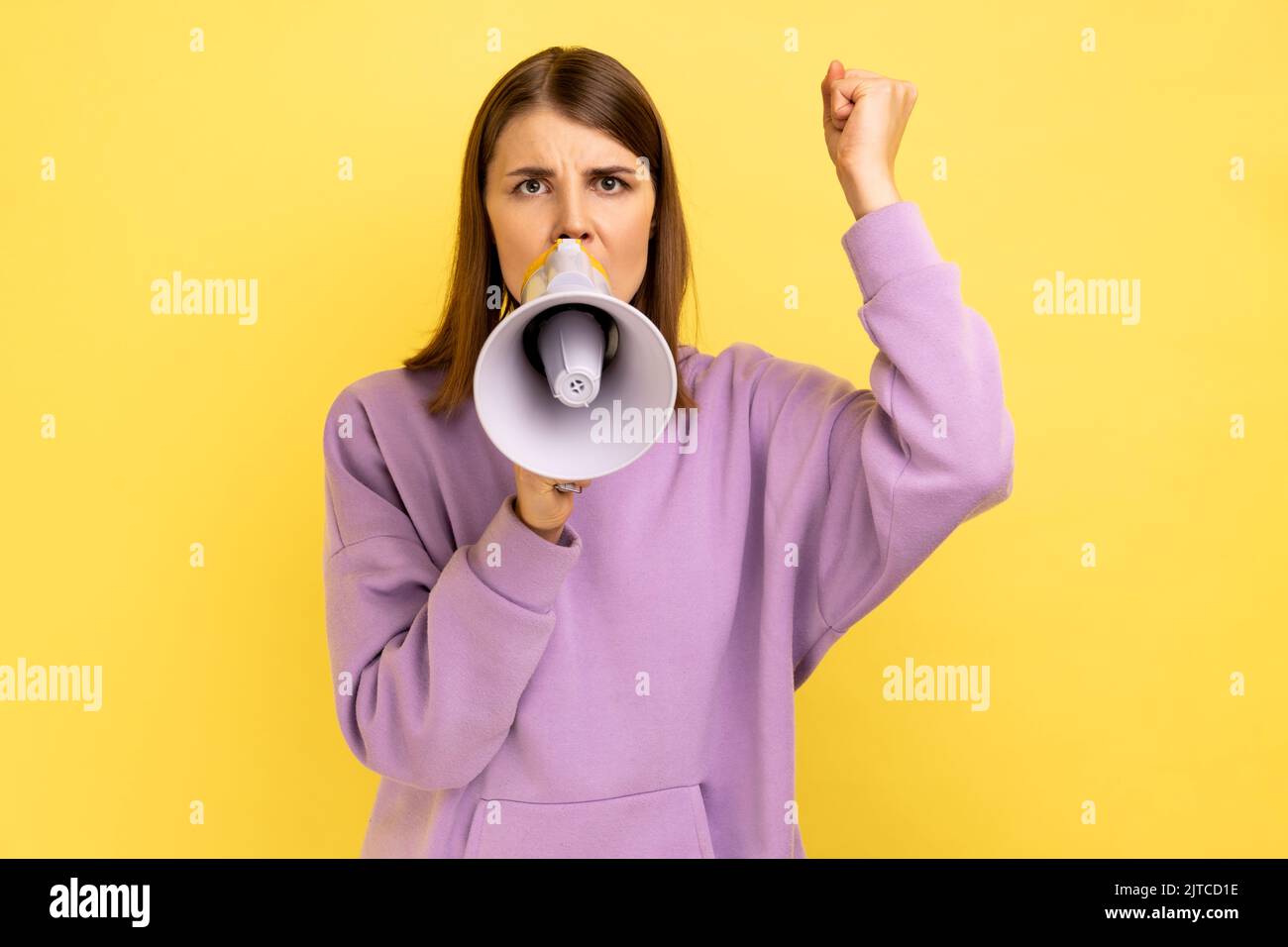 Portrait of woman holding megaphone near mouth, loudly speaking, screaming, making announcement with raised arm, protesting, wearing purple hoodie. Indoor studio shot isolated on yellow background. Stock Photo