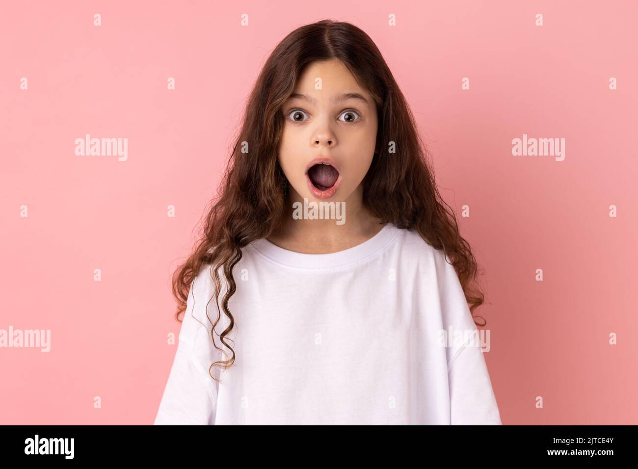 Portrait of surprised astonished little girl wearing white T-shirt looking at camera with open mouth and big eyes, sees something shocked. Indoor studio shot isolated on pink background. Stock Photo