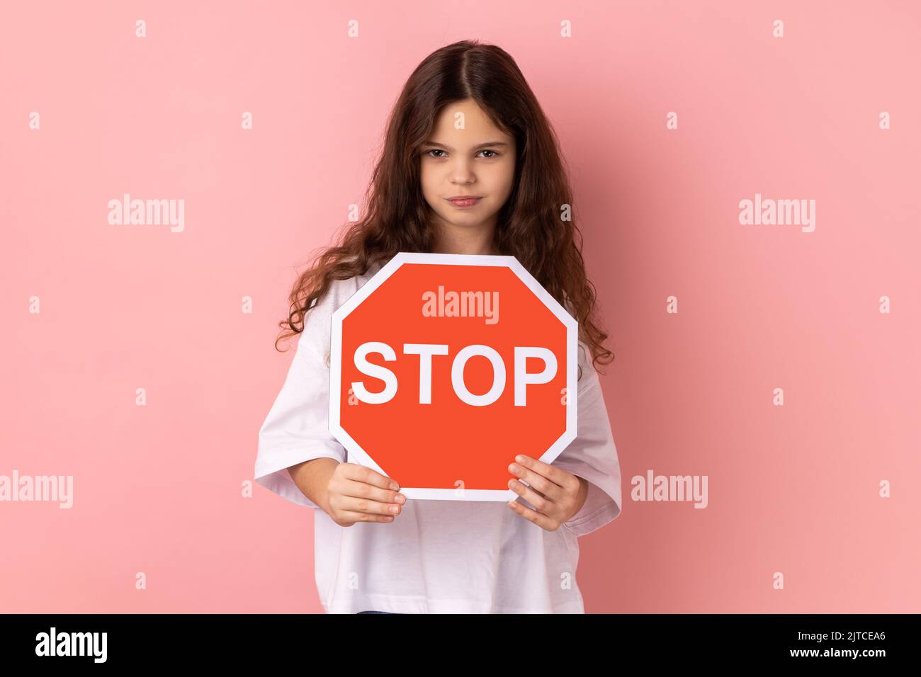 Portrait of strict bossy adorable little girl wearing white T-shirt holding red stop sign looking at camera with serious expression. Indoor studio shot isolated on pink background. Stock Photo