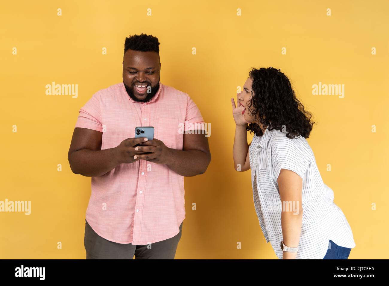 Young couple in casual clothing standing together, woman desperately screaming near happy man using phone and ignoring her, attracting his attention. Indoor studio shot isolated on yellow background. Stock Photo