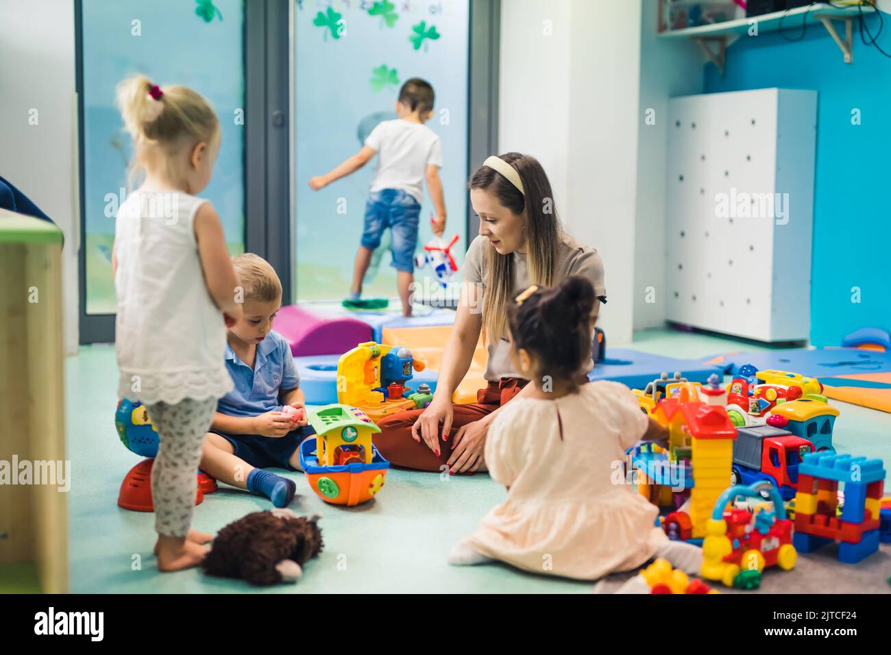Toddlers and their nursery tutor playing with different colorful plastic toys and building blocks while sitting on the floor in a playroom. Concentration, mathematical ability, fine motor and gross motor skills development. . High quality photo Stock Photo