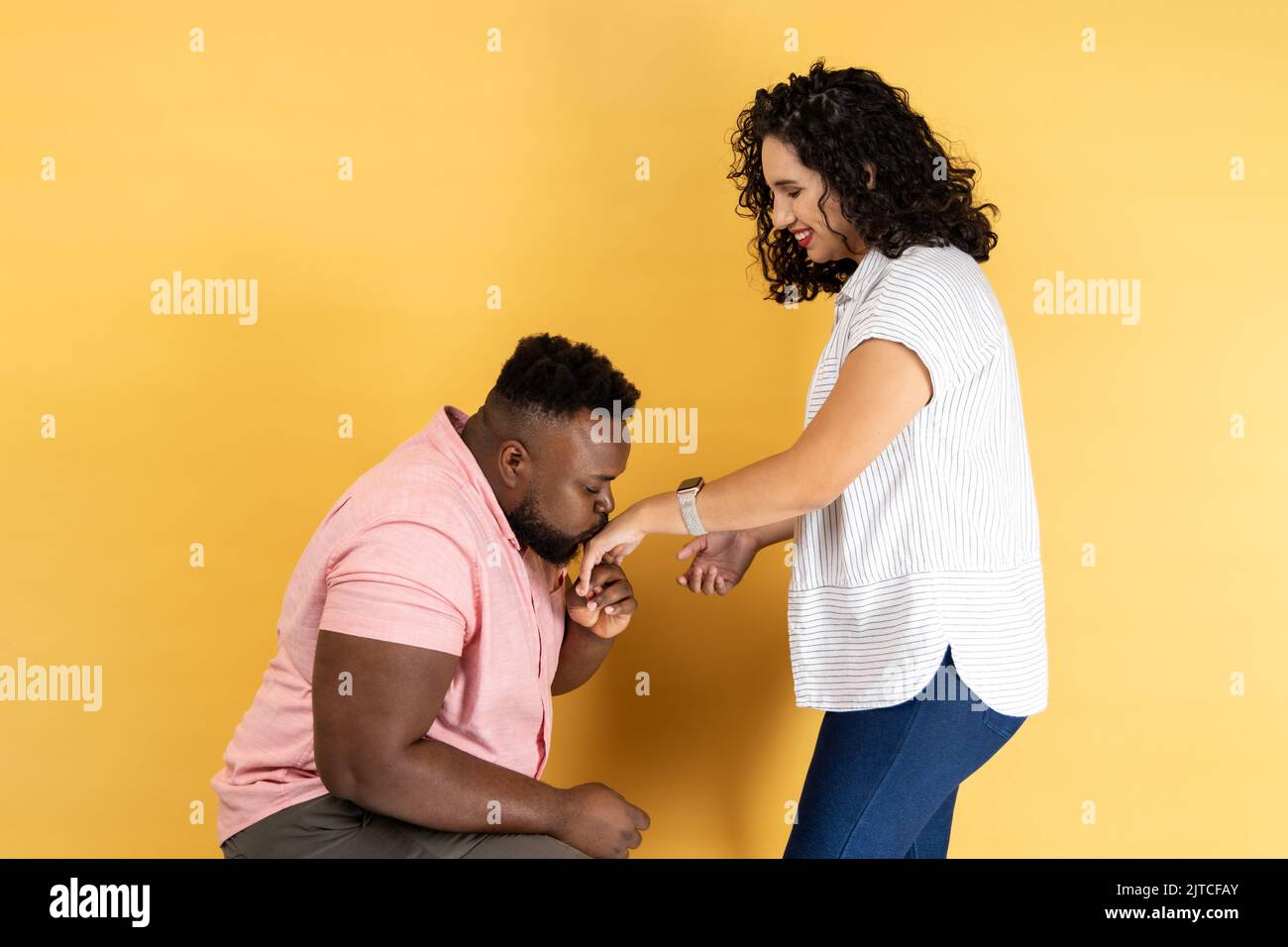 Portrait of romantic young couple in casual clothing posing together, man standing on his knee and kissing her wife hand, expressing love. Indoor studio shot isolated on yellow background. Stock Photo