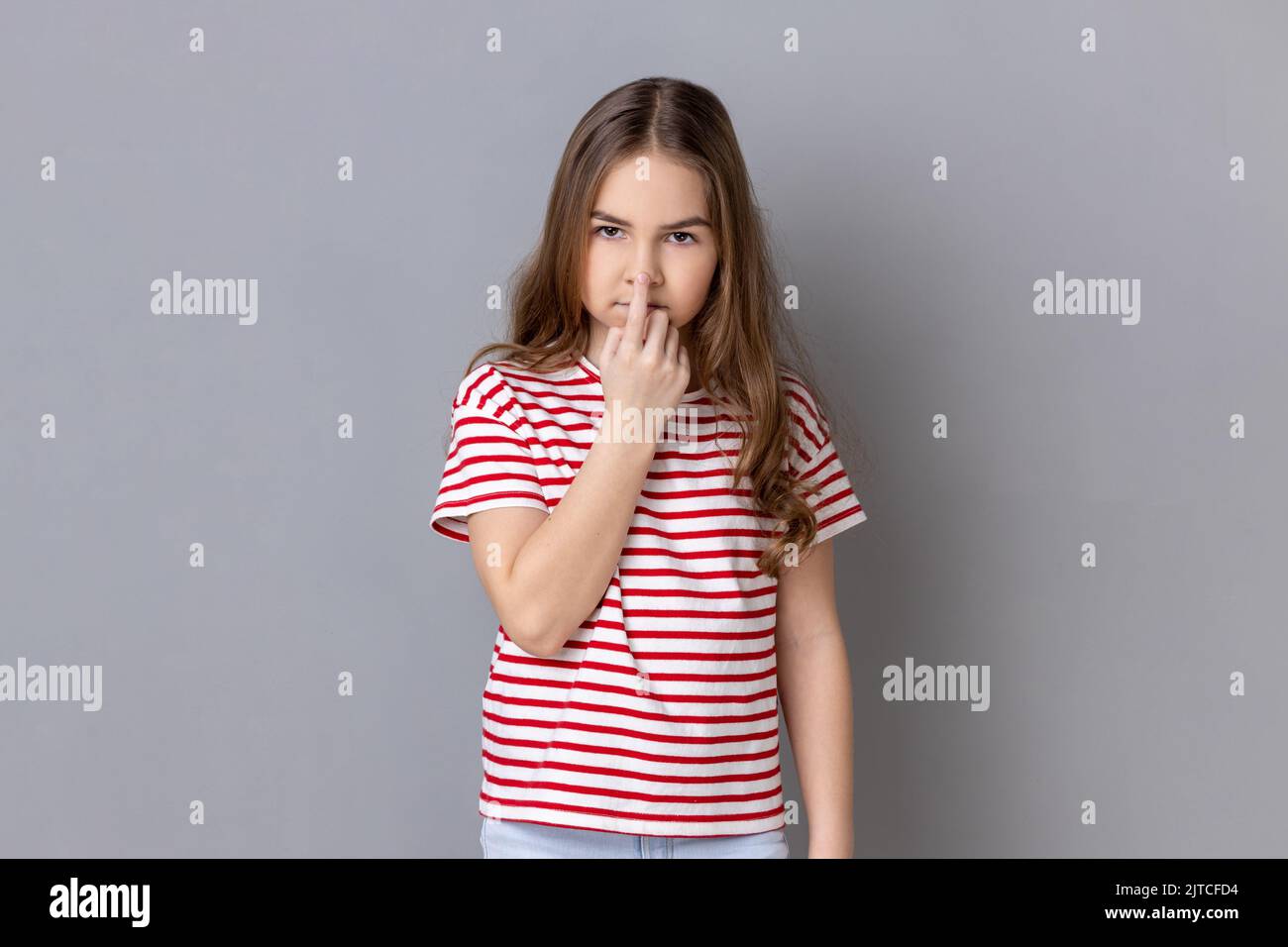 Don't lie. Little girl wearing striped T-shirt looking with incredulous suspicious gaze and touching nose, gesturing you are liar, suspecting falsehood. Indoor studio shot isolated on gray background. Stock Photo