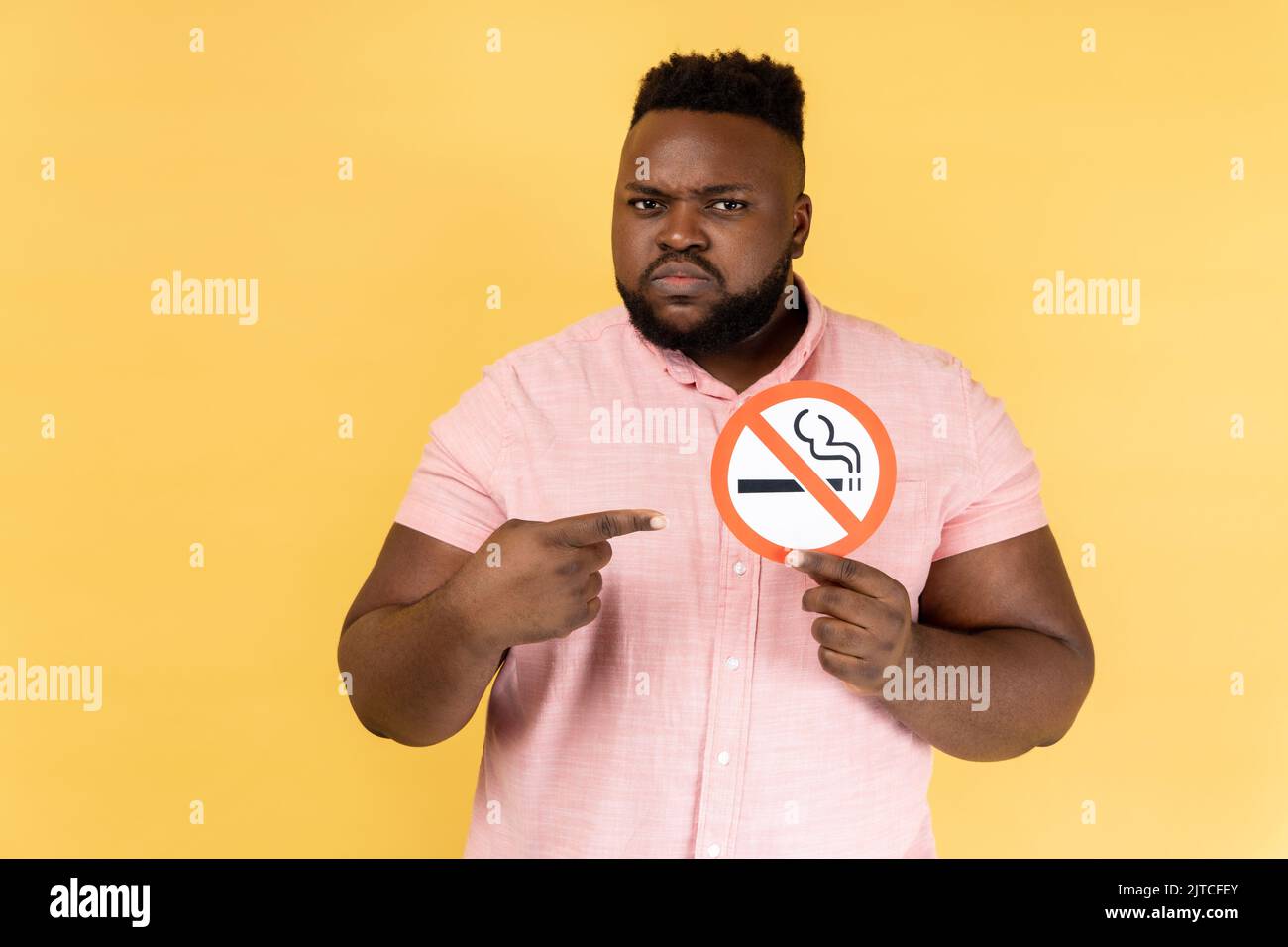 Portrait of serious man wearing pink shirt holding and pointing index finger at no smoking sign in hands, quit smoking, no tobacco day. Indoor studio shot isolated on yellow background. Stock Photo