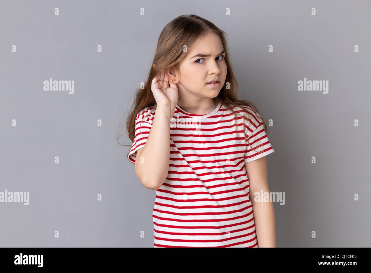 Portrait of little girl wearing striped T-shirt holding hand near ear and listening carefully, having hearing problems, deafness in communication. Indoor studio shot isolated on gray background. Stock Photo
