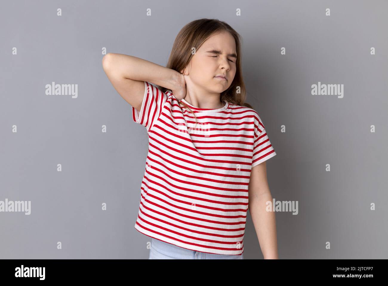 Portrait of unhealthy sick dark haired little girl wearing striped T-shirt standing massaging neck to relieve pain, muscle strain in back. Indoor studio shot isolated on gray background. Stock Photo