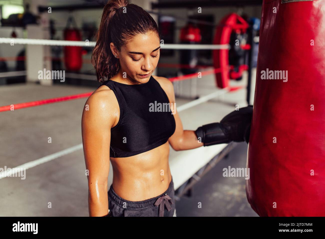 Sweaty female boxer standing next to a punching bag while taking a break. Sporty young woman training in a boxing gym. Stock Photo