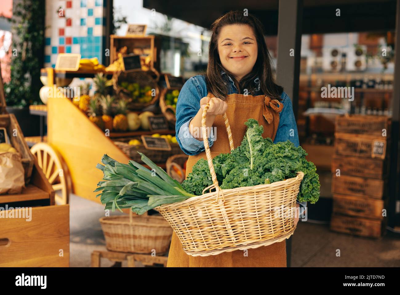 Supermarket employee with Down syndrome holding a basket of fresh organic vegetables in a grocery store. Happy woman with an intellectual disability w Stock Photo