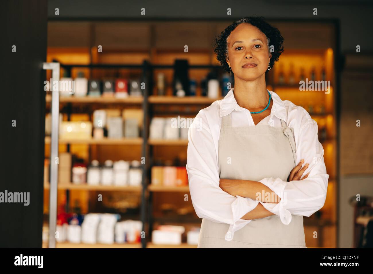 Confident small business owner looking at the camera while standing at the entrance of her grocery store with her arms crossed. Mature entrepreneur ru Stock Photo