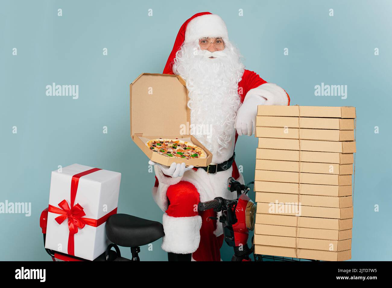 Pizza delivery man dressed as santa claus with an open pizza box in his hand cloaks himself on a stack of boxes on the background of a red scooter and Stock Photo