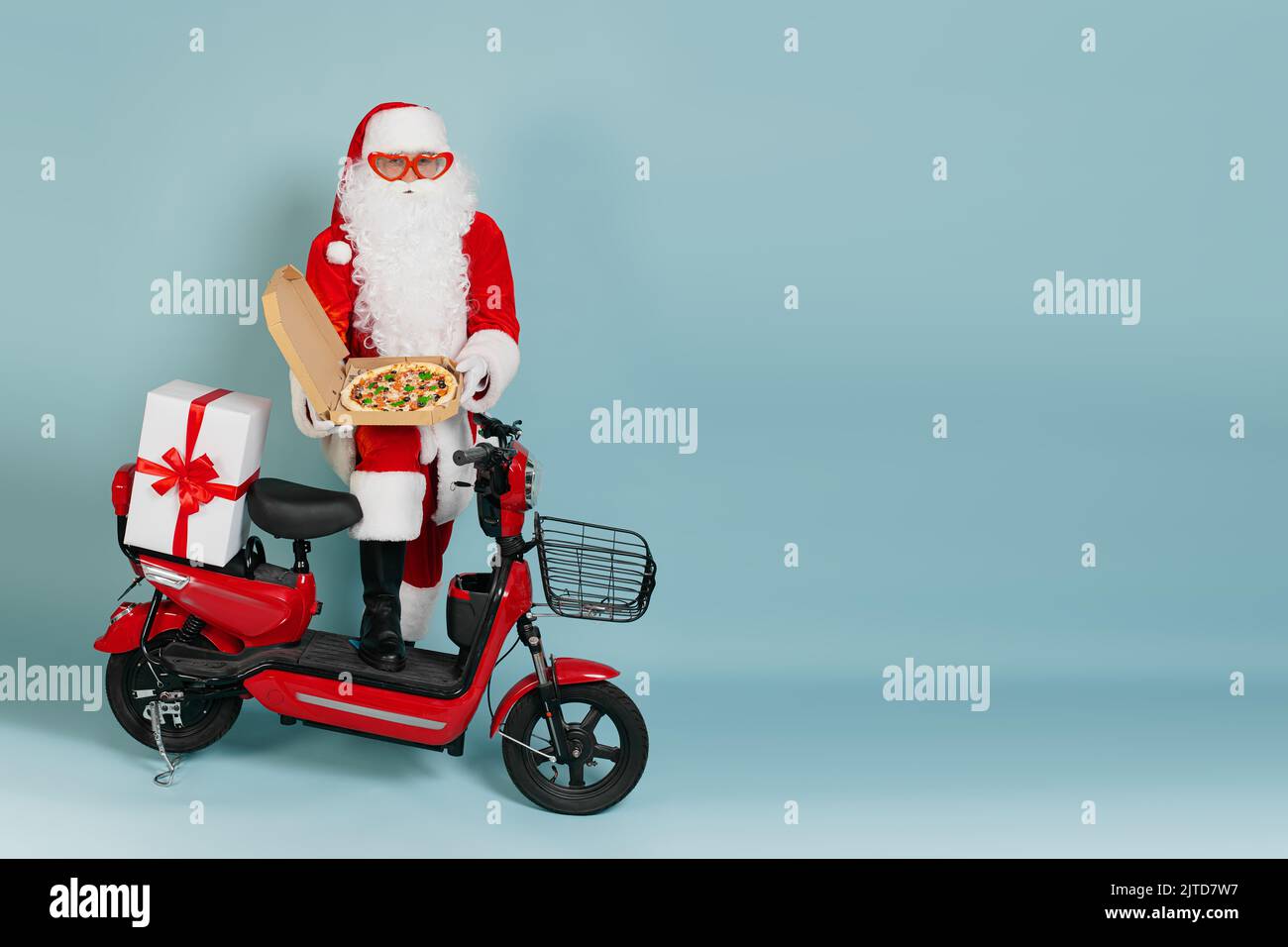 Full length portrait of santa claus in big red heart shaped glasses with an open box of pizza held in his hands in white gloves, against a red moped o Stock Photo