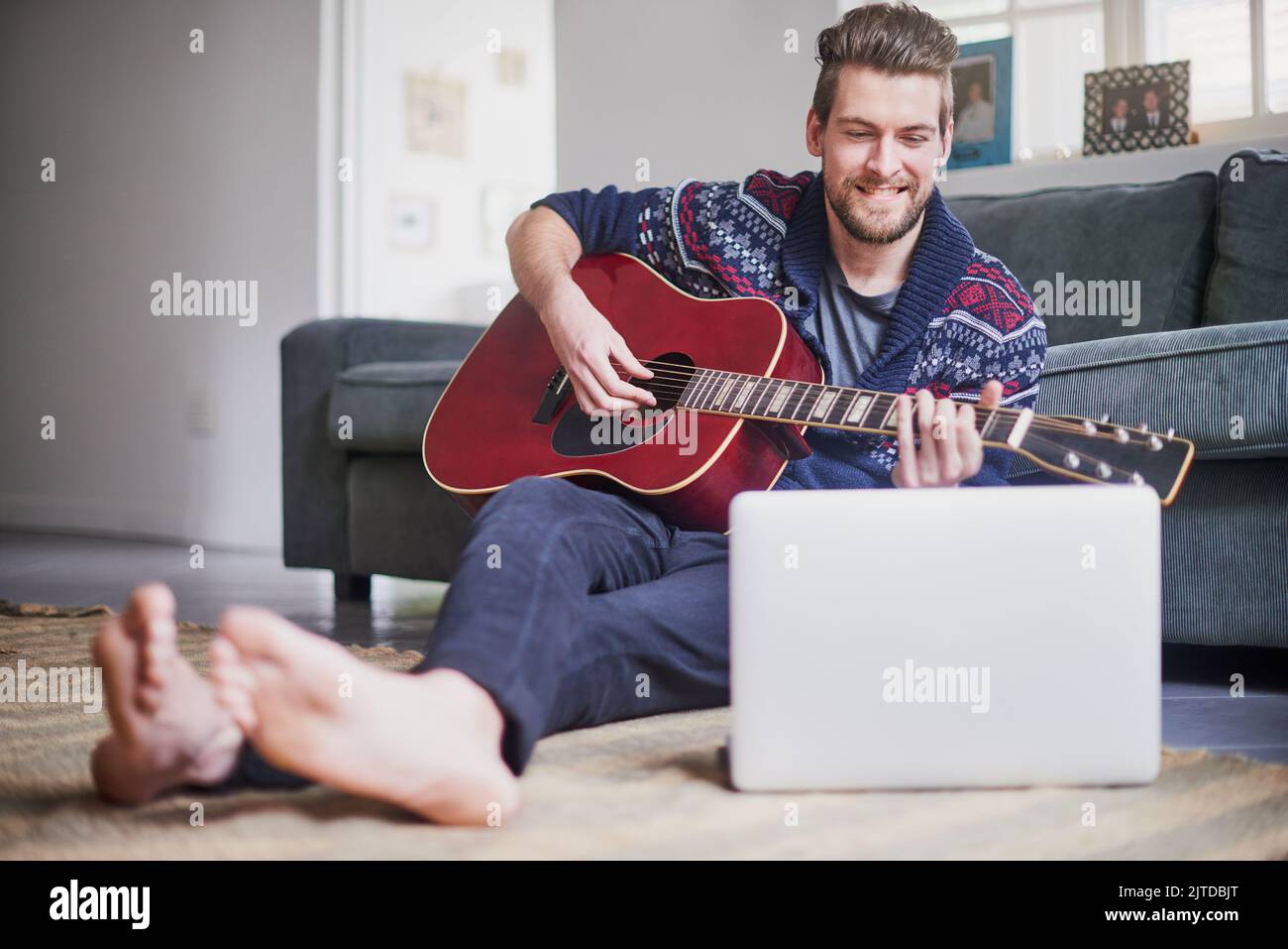 Hes found an easy tutorial to follow online. a handsome young man playing a guitar while watching an online tutorial on his laptop. Stock Photo