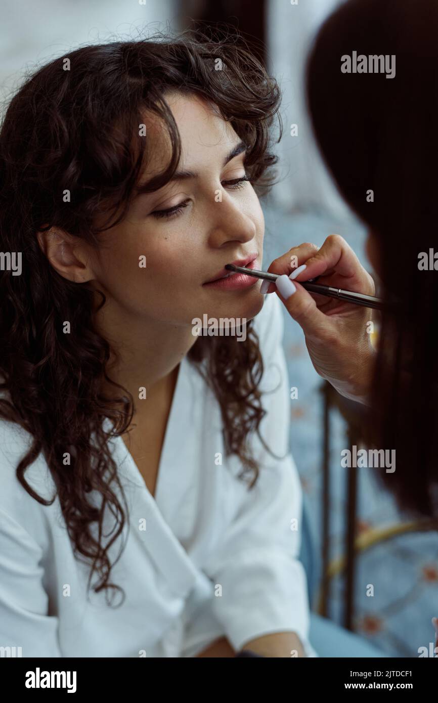 Hand of female makeup artist applying lipstick or gloss on lips of pretty bride with dark long wavy hair on wedding morning Stock Photo