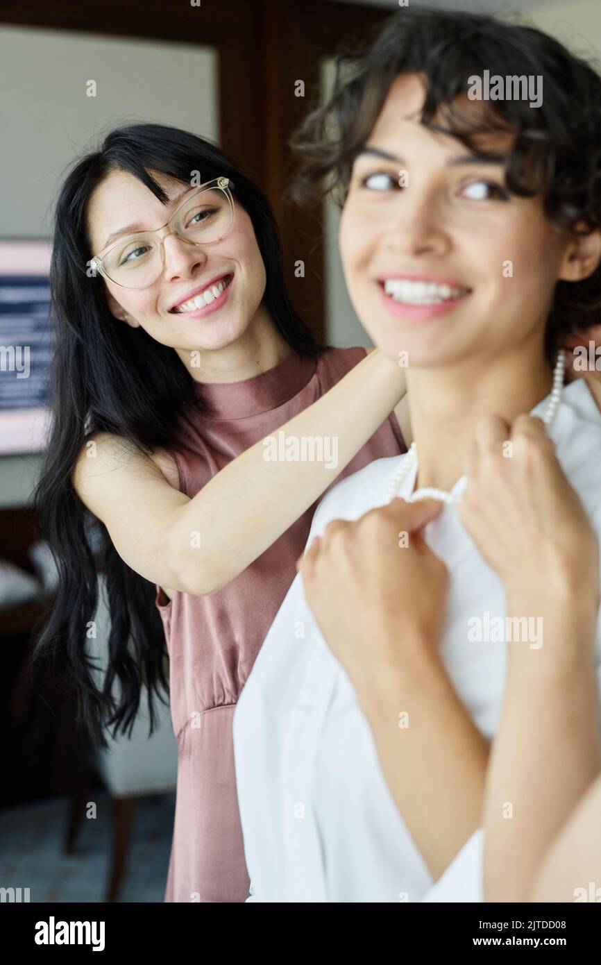 Happy young brunette woman helping her sister in white silk bathrobe put pearl necklace on neck during preparation for wedding Stock Photo