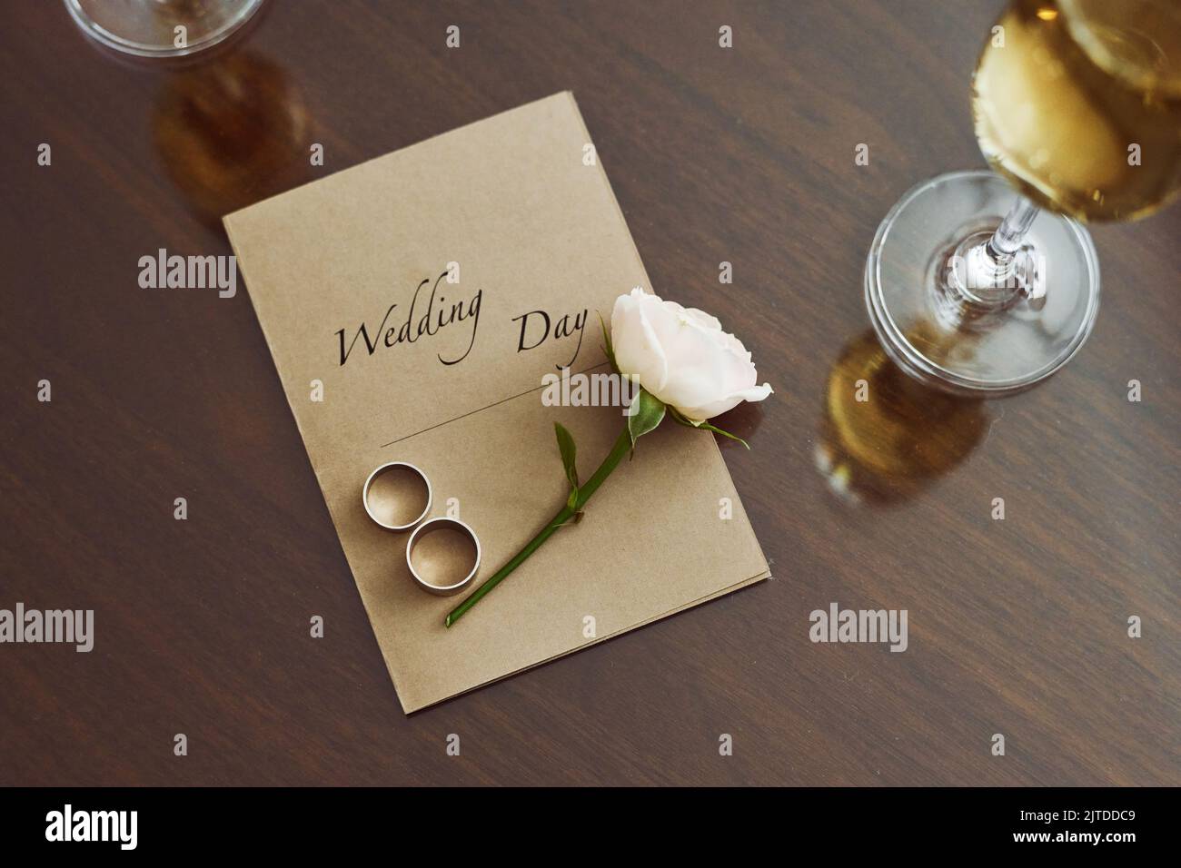 Top view of wedding composition on table consisting of invitation, fresh white rose and two rings surrounded by flutes of champagne Stock Photo