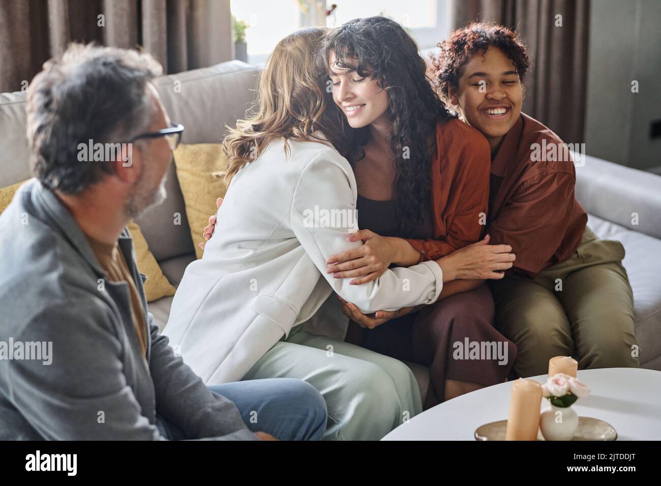 Happy young brunette woman giving hug to mother while sitting on couch next to her cheerful African American girlfriend Stock Photo