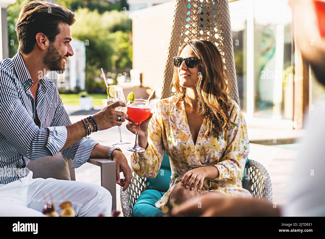A young millennial generation couple celebrates a toast sitting in the resort garden clinking glasses with imaginative cocktails - backyard recreation Stock Photo