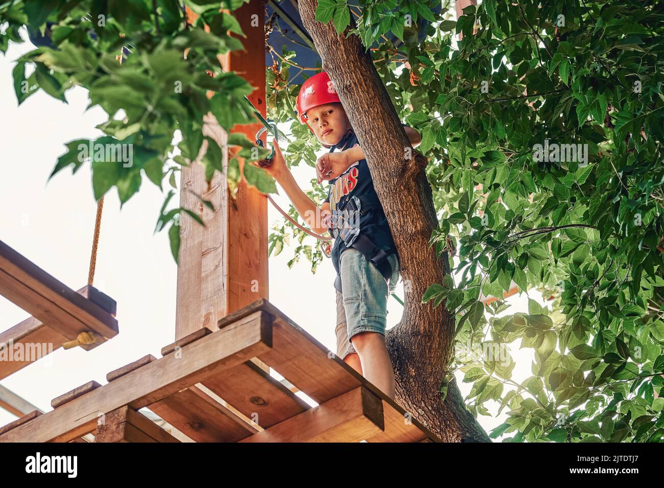 Boy in a helmet and climbing equipment standing on a wooden platform against foliage background, spending time in rope park in summer evening Stock Photo