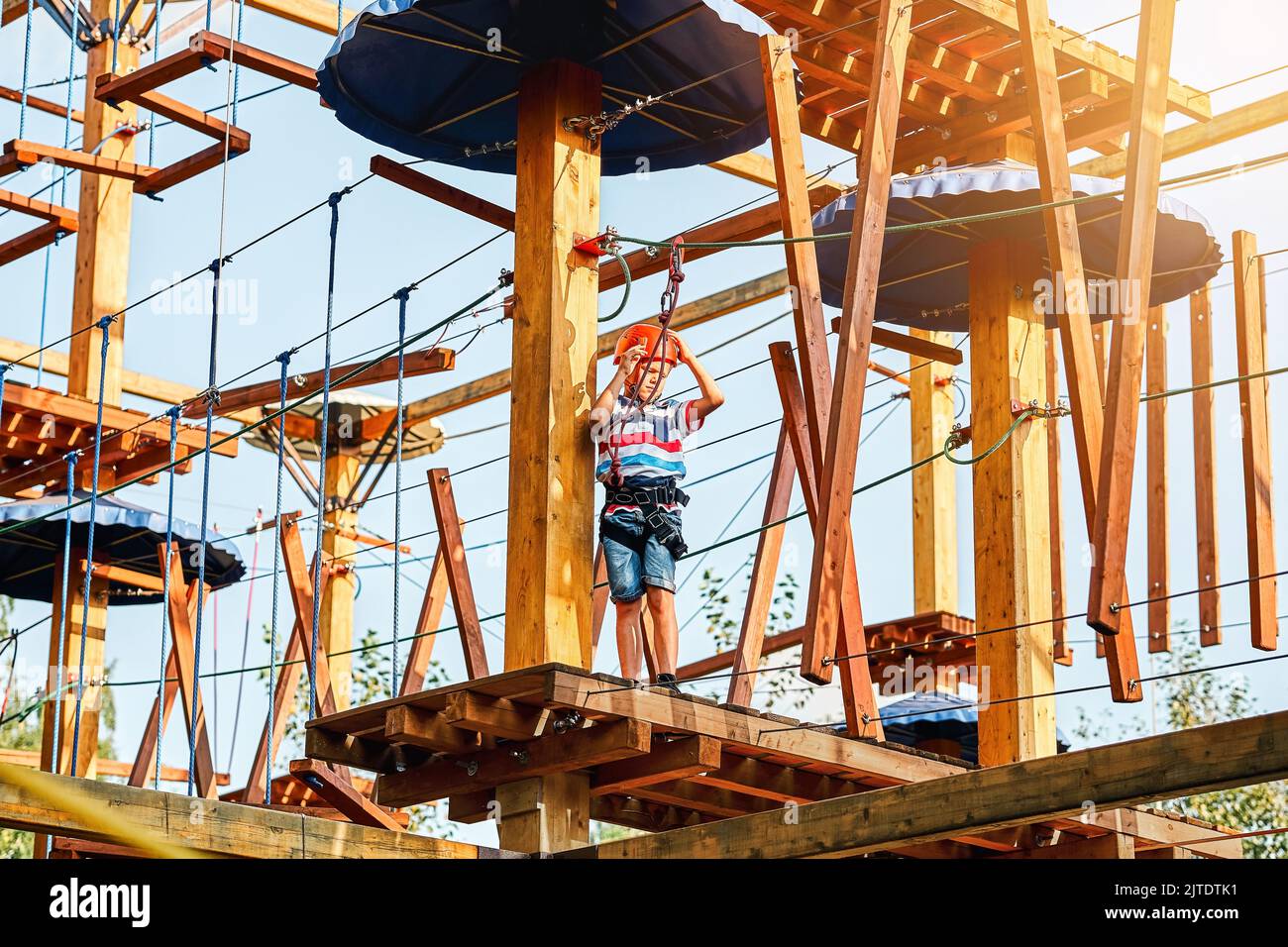 Boy in a helmet and climbing equipment standing on a wooden platform against the sky, spending time in rope park in summer evening Stock Photo