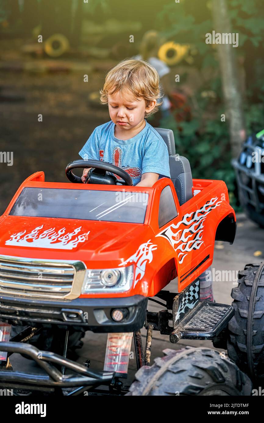 Frustrated toddler boy in blue t-shirt sits in red children car with big wheels in public park in summer evening Stock Photo