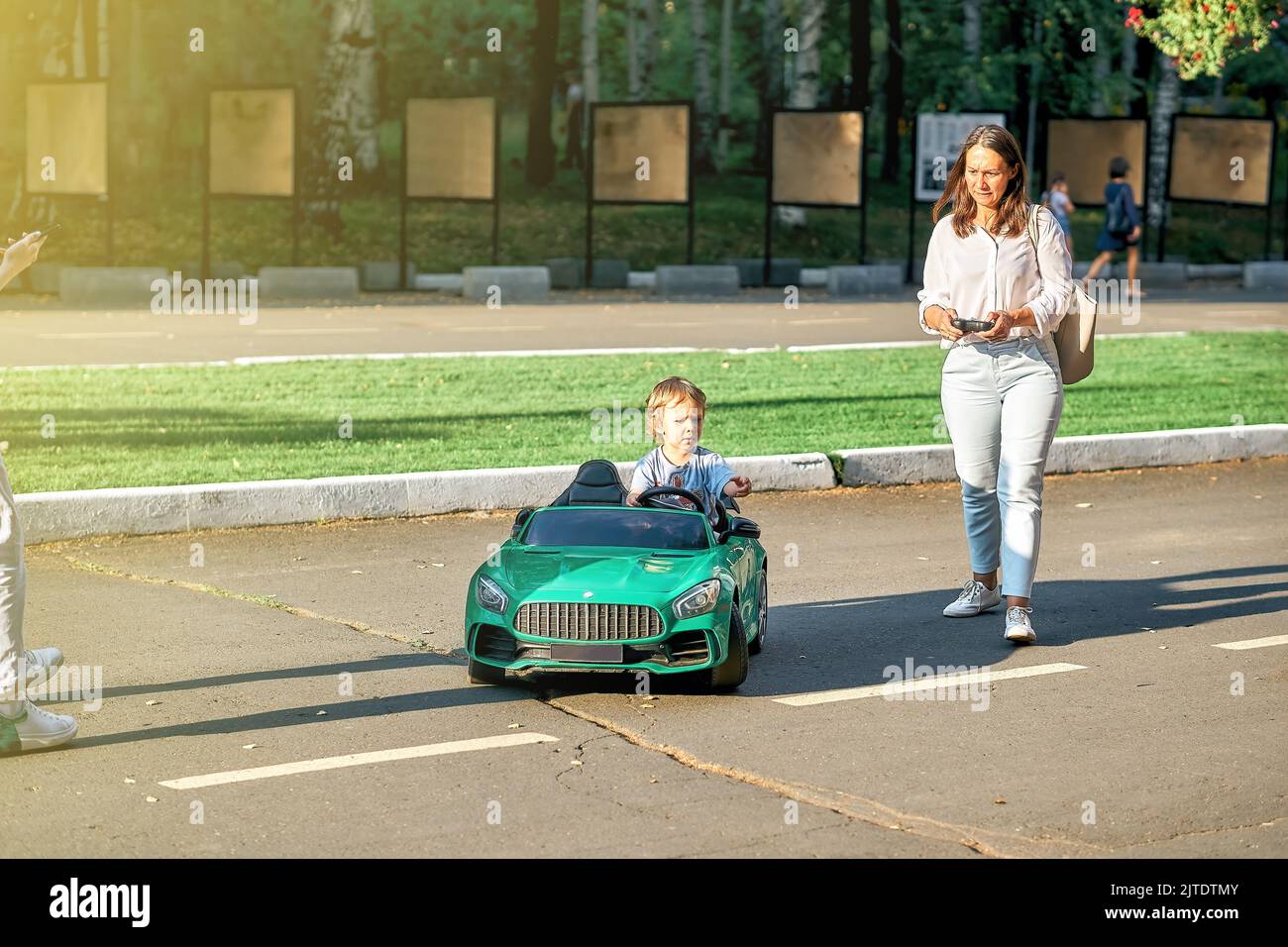 Mom controls children car with remote at walk with toddler child in public park. Toddler boy sits in green car holding steering wheel looking aside Stock Photo