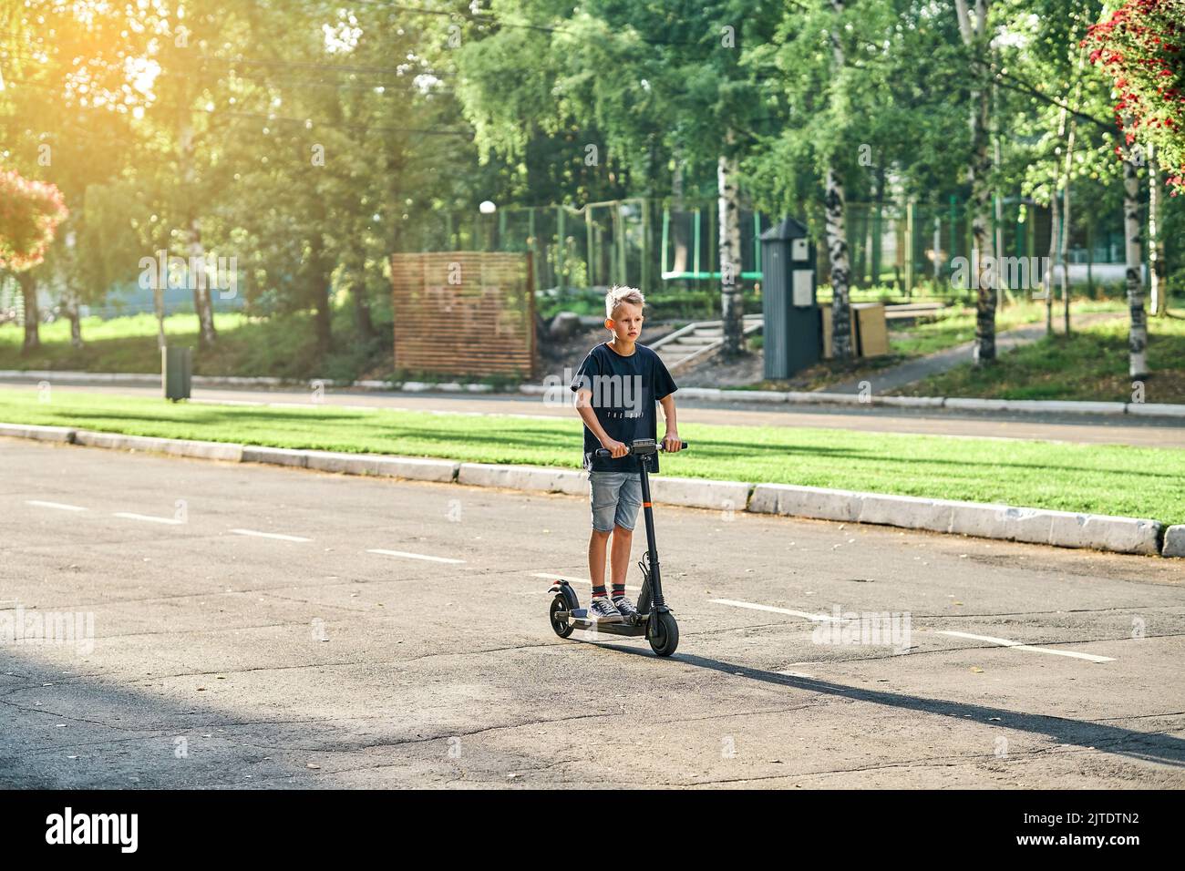 Schoolboy rides electric scooter on road in empty summer park. Child in black t-shirt and denim shorts rides on scooter along marked asphalt road Stock Photo