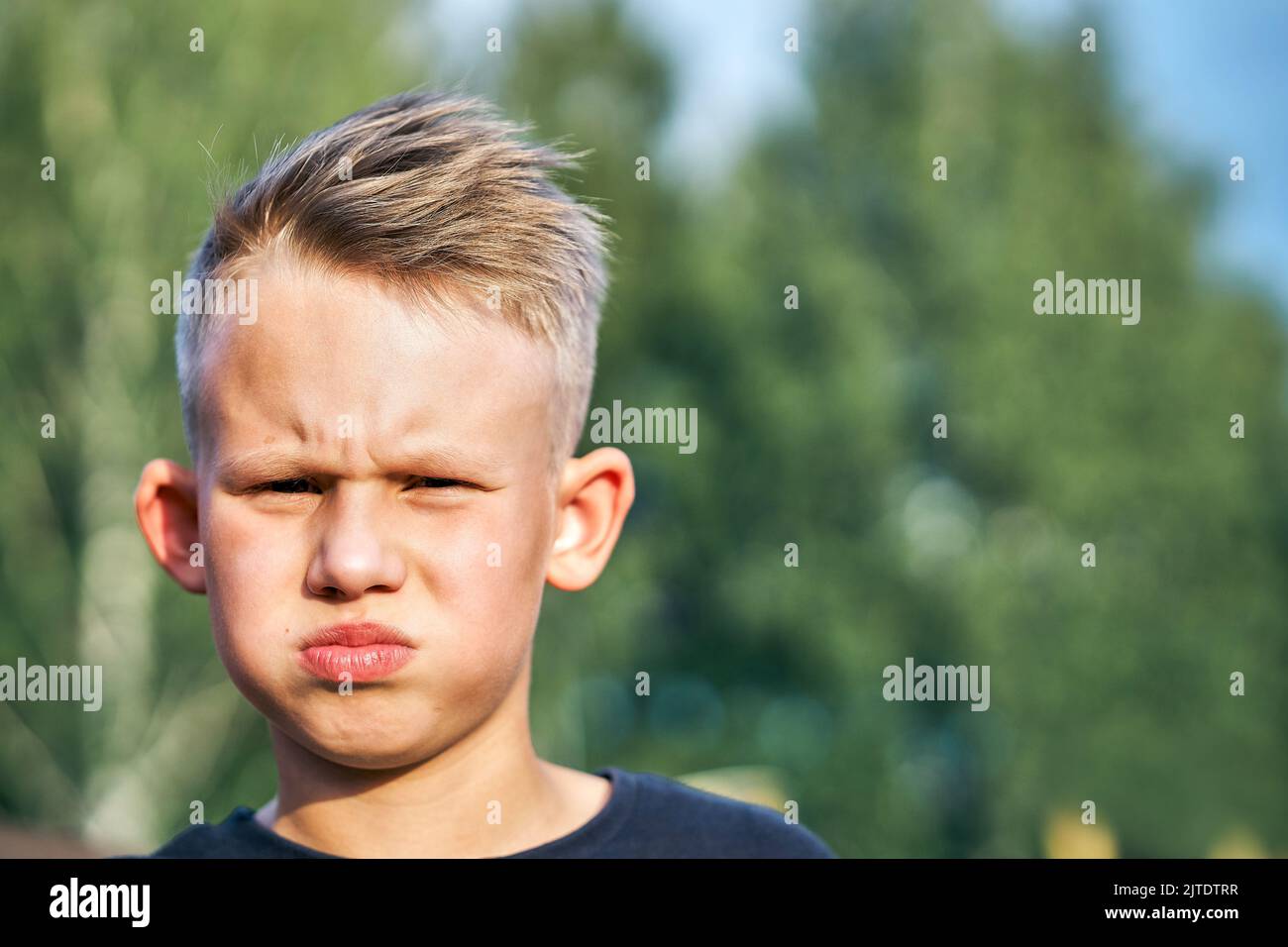 Frustrated little schoolboy with short hairstyle posing for camera on blurred green sunny summer city park extreme closeup Stock Photo