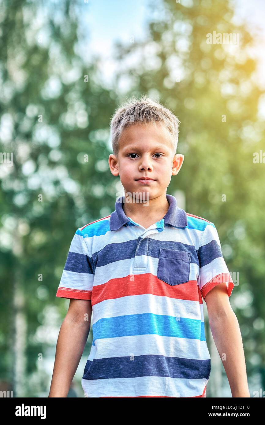 Cute blond boy with short hairstyle in stylish casual clothes posing for camera in green sunny summer city park Stock Photo