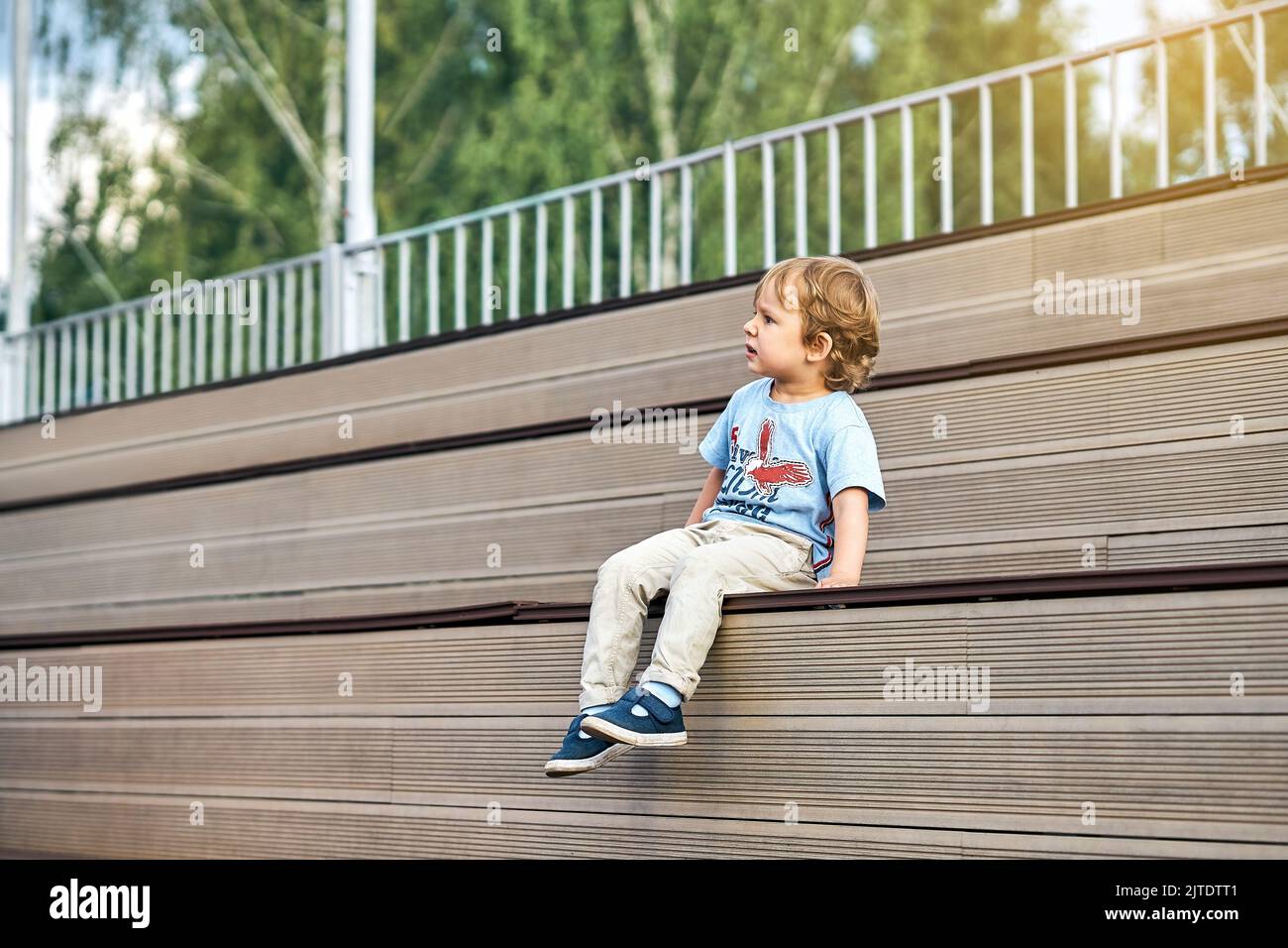 Adorable little boy with funny grimace in stylish casual clothes sit on wooden bench watching movie on huge screen in public park Stock Photo