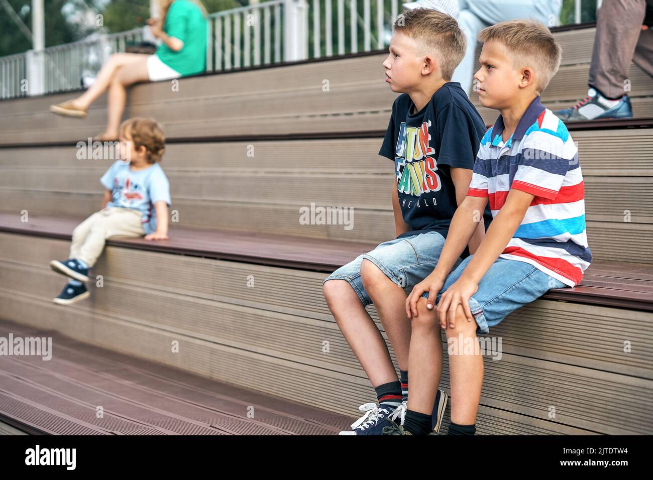 Boys in stylish casual clothes sit on wooden bench watching movie on huge screen in public park Stock Photo