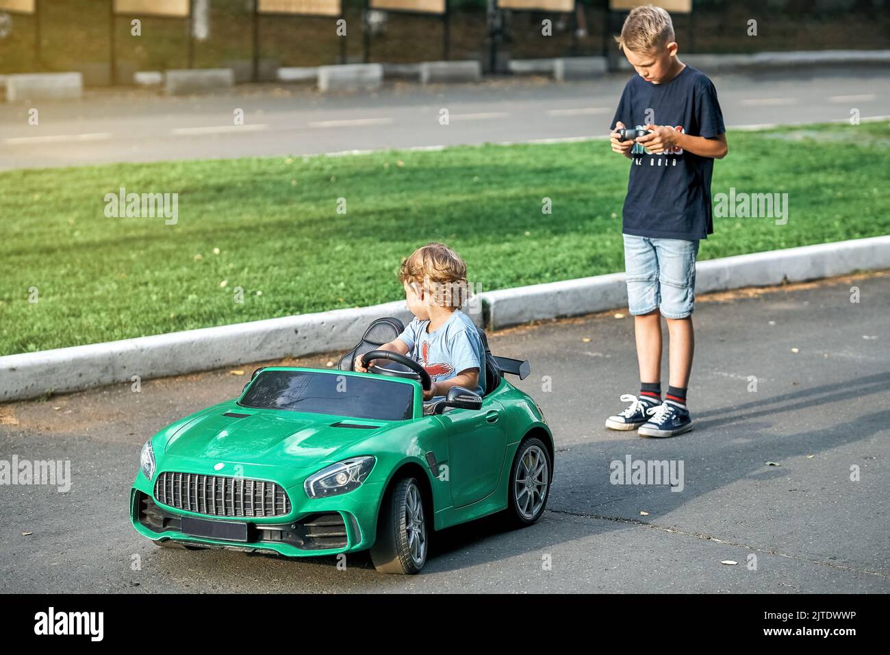 Elder brother controls children car with remote at walk with little brother in public park. Toddler boy sits in green car holding steering wheel Stock Photo