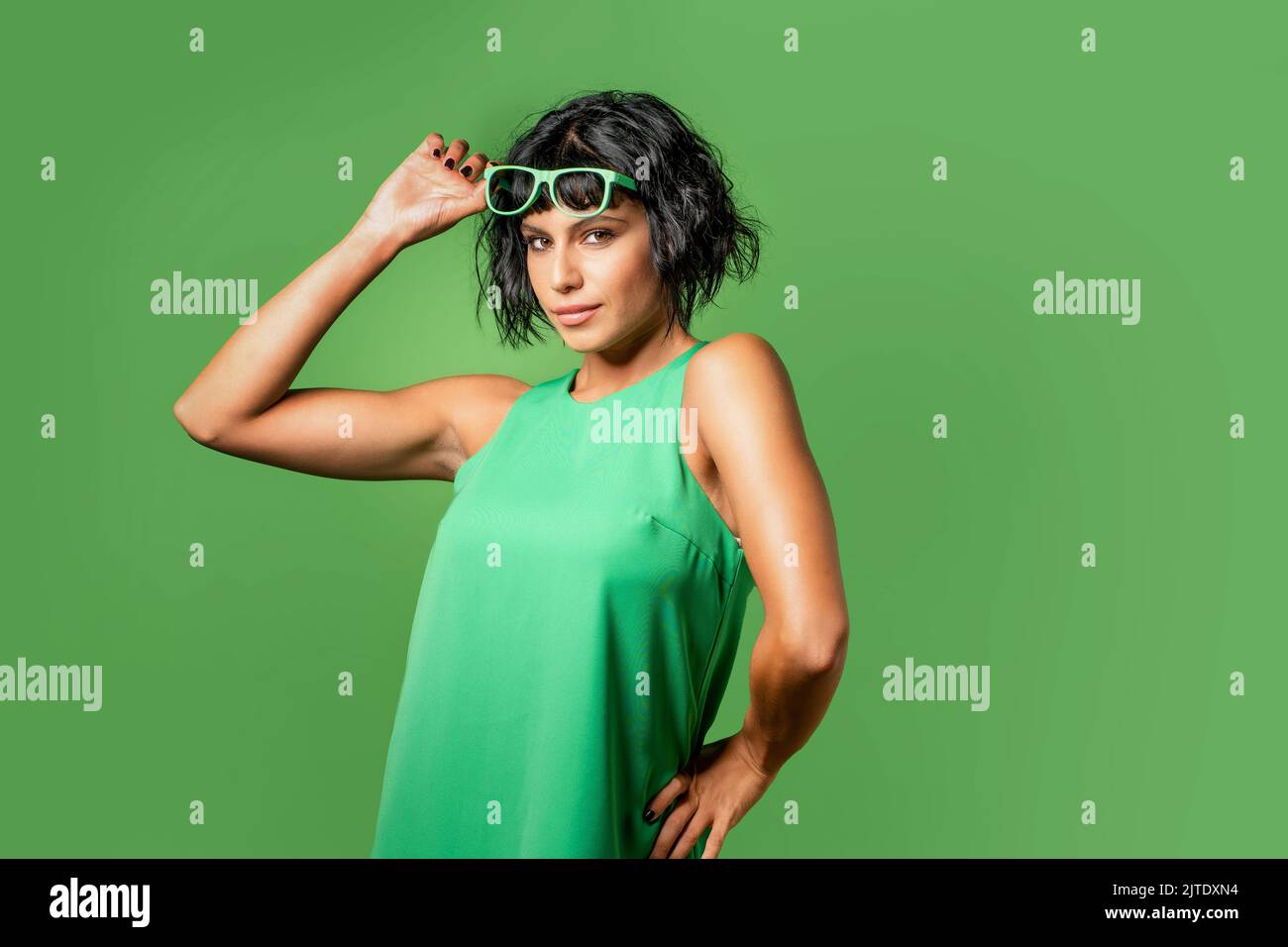 Winking young brunette woman 20s years old wears tank top touching raise sunglasses up - isolated on plain vivid green background - studio portrait - Stock Photo